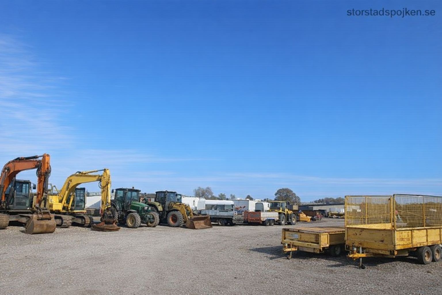 En samling tunga maskiner och släpvagnar på en grusplan under klarblå himmel.