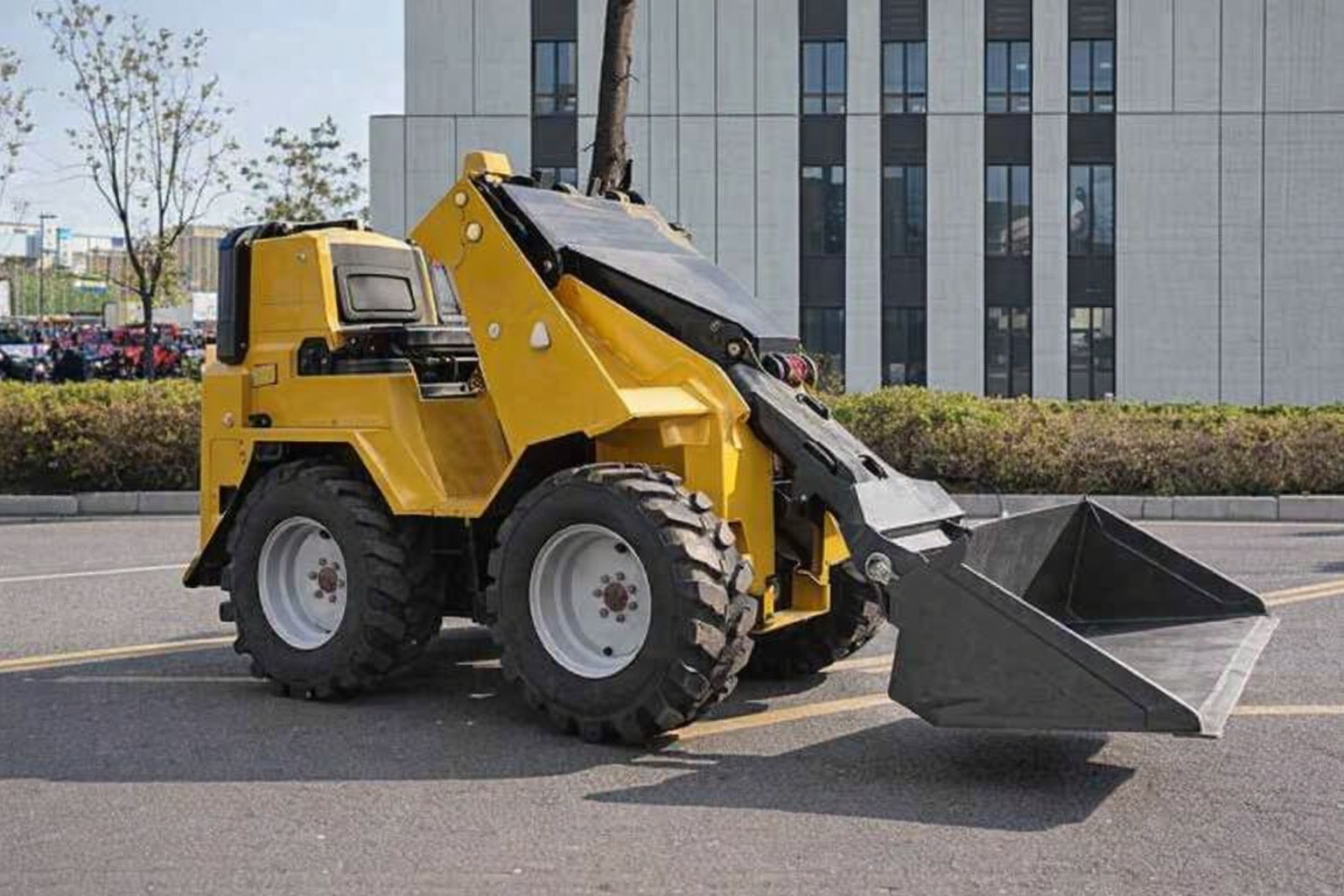 En gul kompakt hjullastare parkerad på en asfalterad yta framför en kontorsbyggnad.